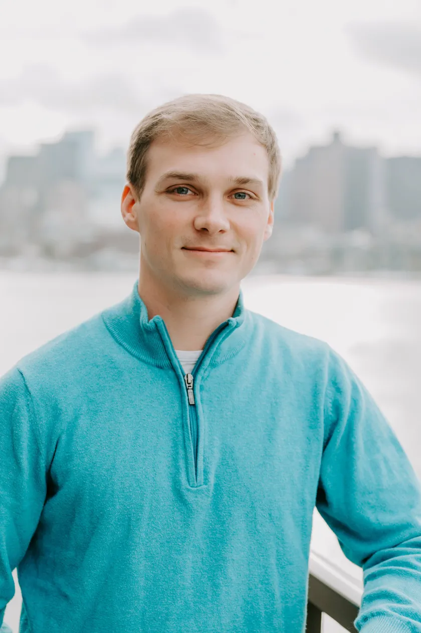 Greg Ellson in a teal quarter-zip, smiling, with a soft city skyline behind him.