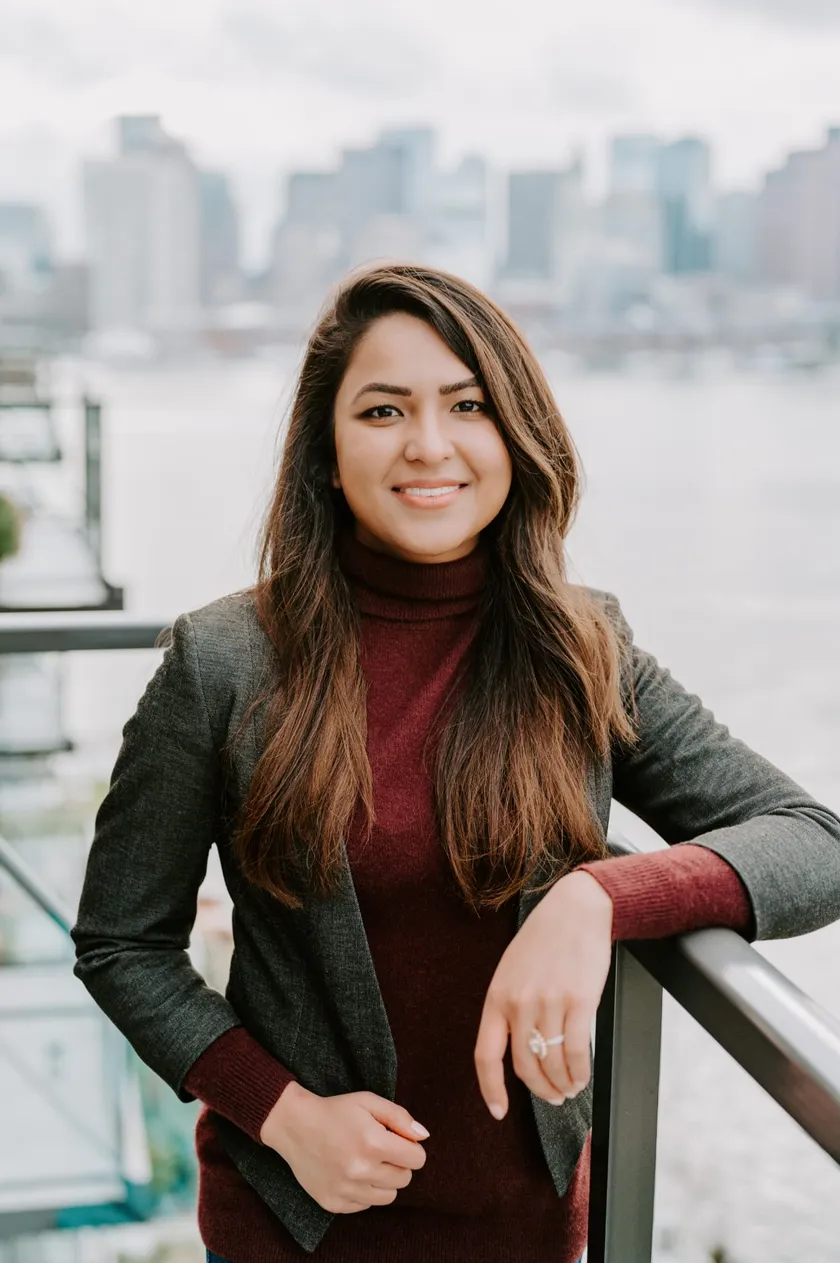 Srishti Ellson in a grey blazer and burgundy turtleneck, smiling, with a soft city skyline behind her.
